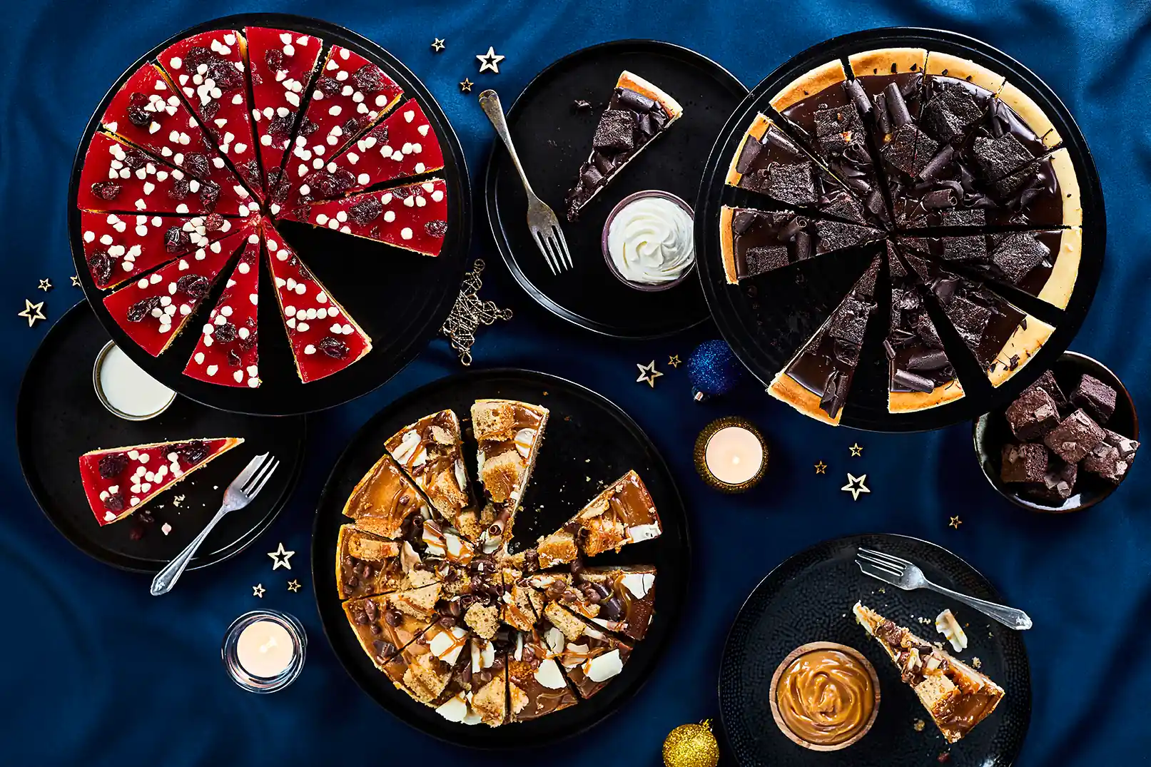 Overhead view of a festive dessert spread on a deep blue tablecloth, featuring sliced cheesecakes and tarts on black plates: a red berry cheesecake with white chocolate dots, a dark chocolate cheesecake topped with chocolate shards, and a caramel and biscuit cheesecake. Plates, forks, candles, and gold star decorations are scattered around, with a few slices removed to show the textures.