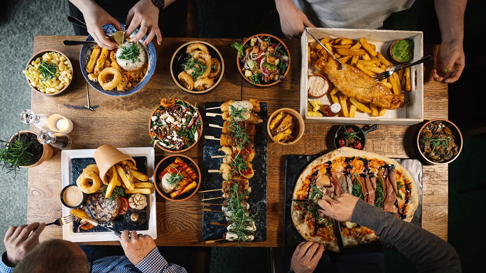 Overhead view of a pub table filled with shared dishes including pizza, fish and chips, skewers, fries, salads and sides, with diners’ hands reaching in to serve and eat together.